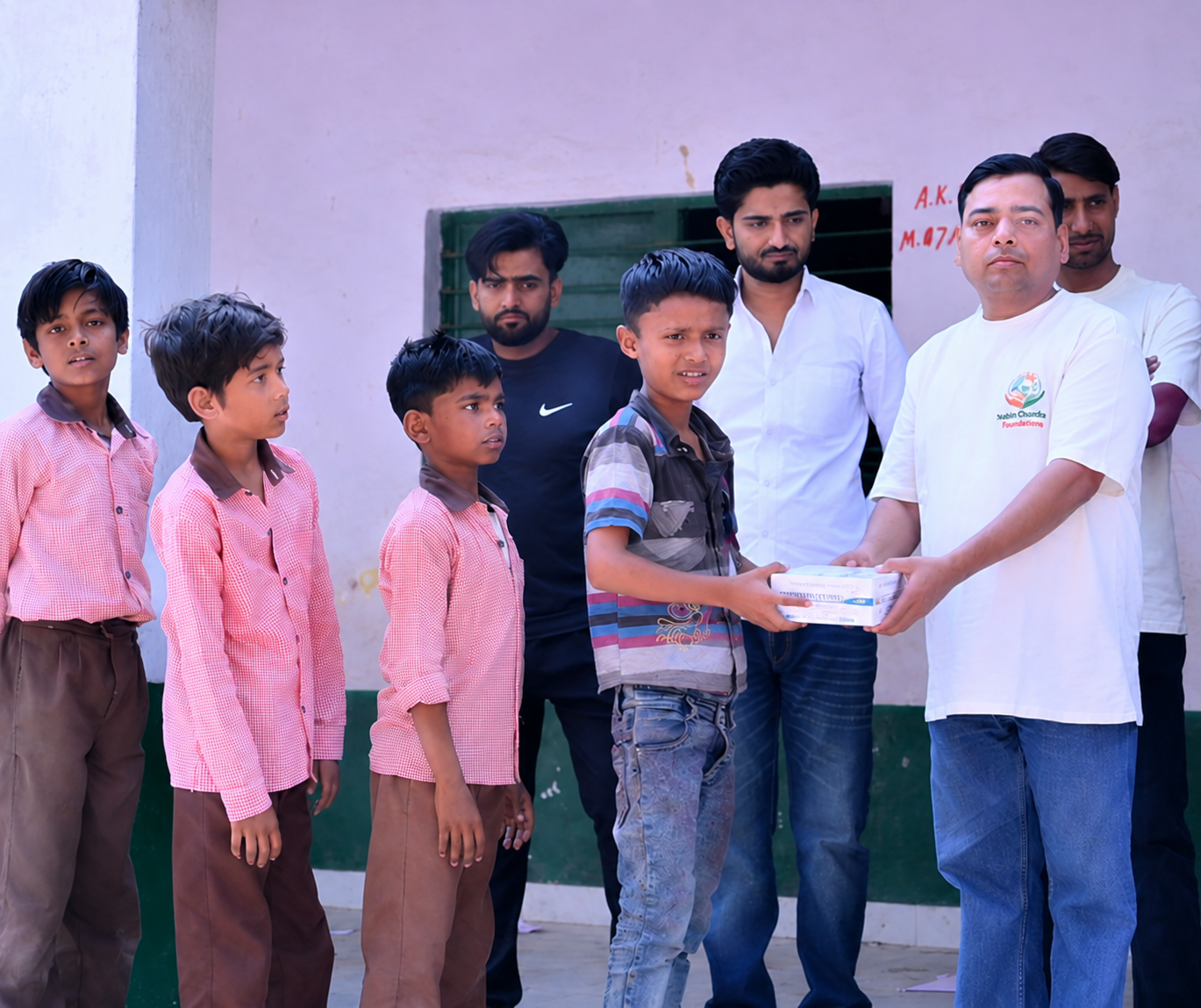 NCF volunteer handing a school supply kit to a boy while uniformed classmates wait outside a school building
